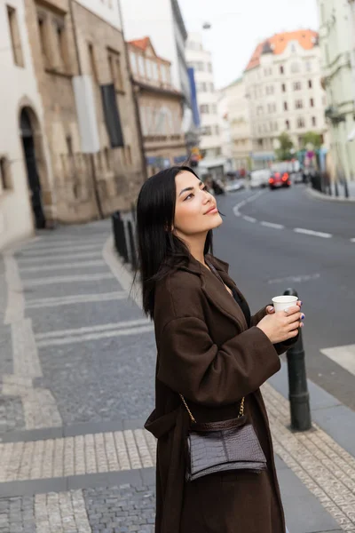Jeune femme brune en manteau avec sac à main tenant tasse en papier près de la route sur la rue urbaine à Prague — Photo de stock
