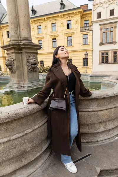 Elegante turista de casaco segurando copo de papel perto da fonte na Praça Charles, em Praga — Fotografia de Stock