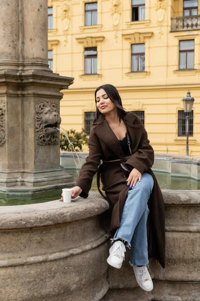 Turista na moda em casaco segurando copo de papel enquanto sentado na fonte na Praça Charles, em Praga — Fotografia de Stock