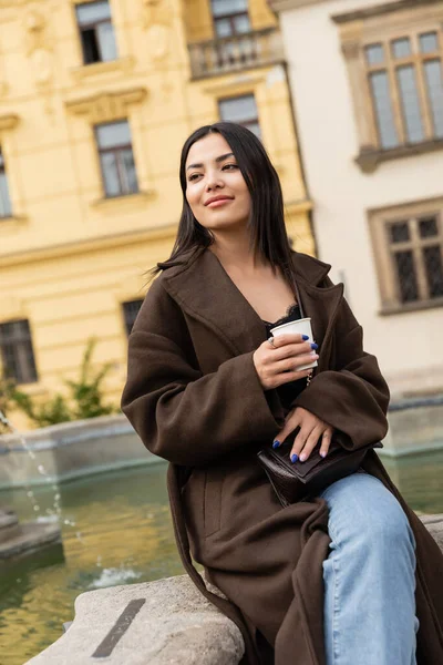 Mujer sonriente con abrigo sosteniendo una taza de papel cerca de la fuente en la Plaza Charles en Praga - foto de stock