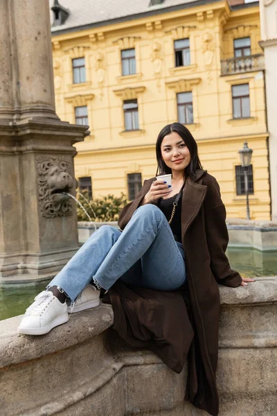 Stylish young woman in coat holding paper cup near old fountain in Charles Square in Prague — Stock Photo