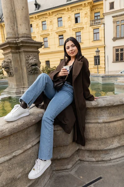Mujer de moda en abrigo sosteniendo taza de papel cerca de la antigua fuente en la Plaza Charles en Praga - foto de stock