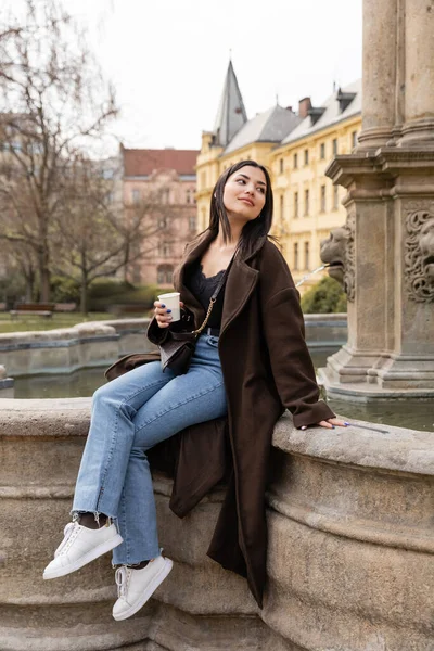 Stylish young tourist in coat holding paper cup near old fountain in Charles Square in Prague — Stock Photo