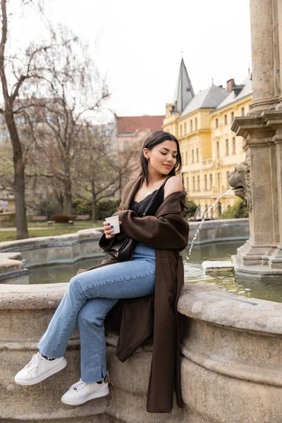 Молодая женщина в пальто и джинсах держит бумажную чашку возле фонтана на Карловой площади в Праге — стоковое фото