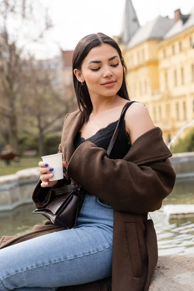 Молодая брюнетка в пальто с бумажной чашкой возле размытого фонтана на открытом воздухе в Праге — стоковое фото