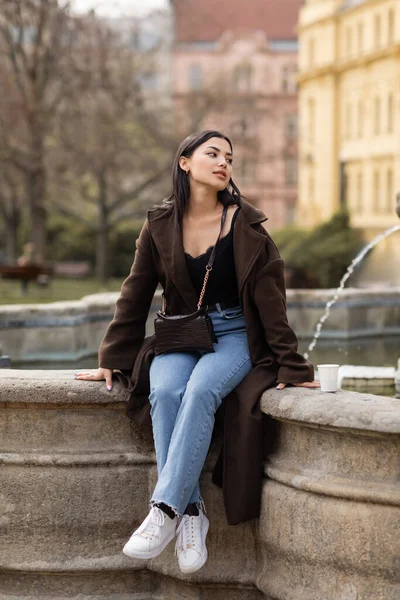 Stylish traveler in coat looking away near paper cup and fountain in Prague — Stock Photo