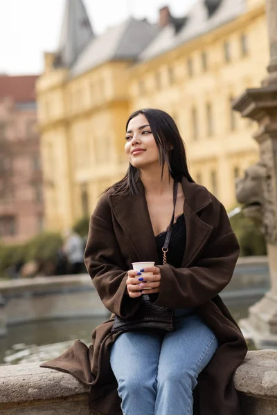 Mulher morena positiva no casaco segurando copo de papel perto de fonte turva na Praça Charles, em Praga — Fotografia de Stock