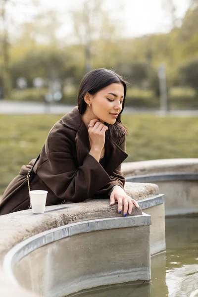 Stylish brunette woman in coat looking at fountain near paper cup on street in Prague — Stock Photo