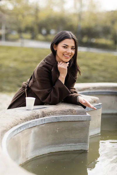 Cheerful tourist in coat looking at camera near paper cup and fountain in Prague — Stock Photo
