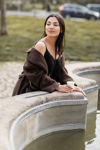 Молода жінка в коричневому пальто тримає паперову чашку біля фонтану в Празі. — стокове фото