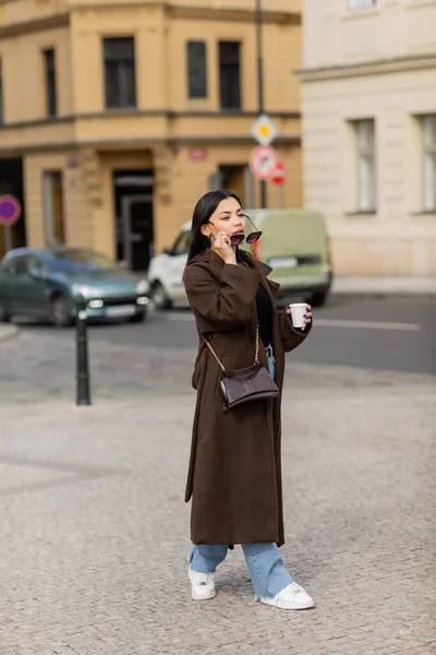 Stylish woman with paper cup holding sunglasses on street in Prague — Stock Photo