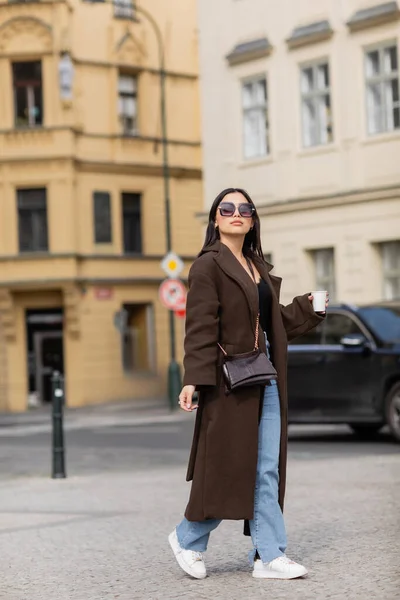 Mujer de moda en abrigo y gafas de sol sosteniendo taza de papel mientras camina por la calle en Praga - foto de stock