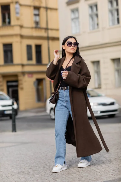 Turista de moda en abrigo y gafas de sol sosteniendo taza de papel mientras camina por la calle en Praga - foto de stock