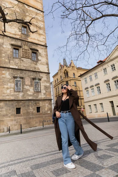 Elegante joven con abrigo y gafas de sol caminando por la calle urbana de Praga - foto de stock