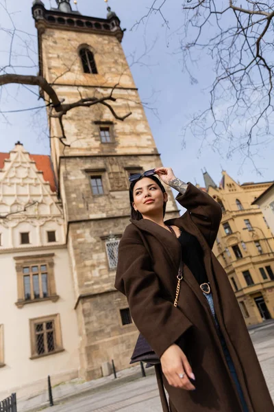 Baixo ângulo vista de mulher tatuada elegante no casaco segurando óculos de sol na rua em Praga — Fotografia de Stock