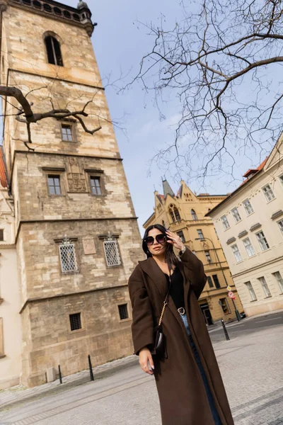 Mulher elegante de casaco e óculos de sol perto de edifícios em Praga — Fotografia de Stock