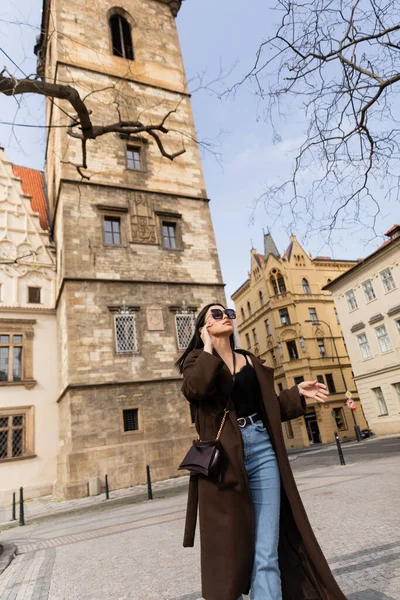 Mujer joven de moda en abrigo y gafas de sol caminando por la calle urbana de Praga - foto de stock