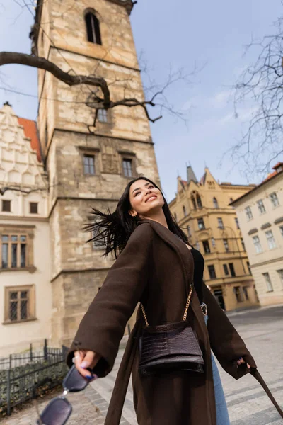 Cheerful woman in coat holding sunglasses on street in Prague — Stock Photo