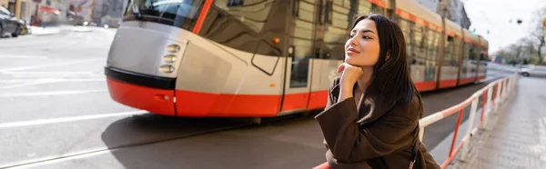 Trendy young woman in coat standing near road with tram on street in Prague, banner — Stock Photo