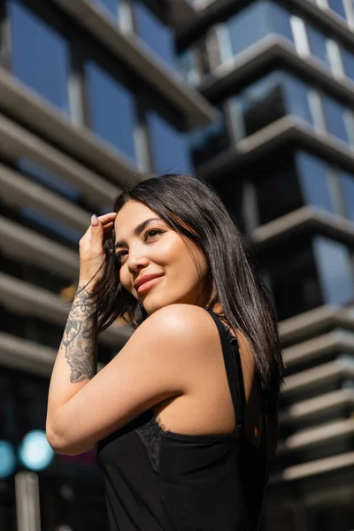 Sorridente donna tatuata che tocca i capelli e distoglie lo sguardo sulla strada urbana — Foto stock