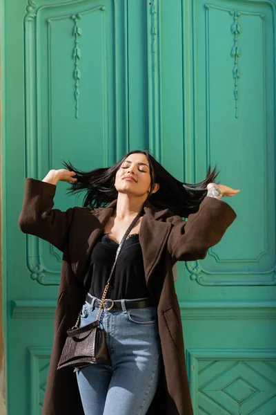 Молода жінка в пальто торкається волосся біля бірюзових дверей будівлі на міській вулиці — стокове фото