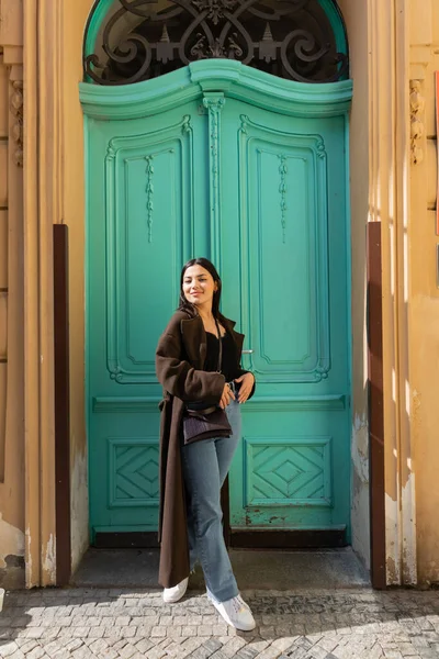 Mujer sonriente en abrigo mirando la cámara cerca de la puerta turquesa del edificio en la calle en Praga - foto de stock
