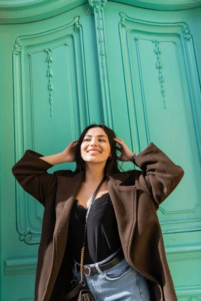 Glückliche junge Frau im Mantel berührt Haare in der Nähe der türkisfarbenen Tür eines Gebäudes auf der Straße — Stockfoto