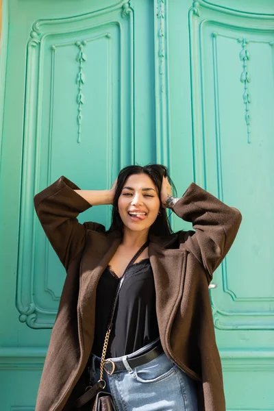 Junge Frau im Mantel streckt vor Kamera vor türkisfarbener Haustür die Zunge heraus — Stockfoto