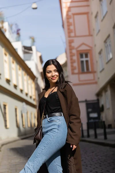 Happy brunette woman in coat and jeans looking away on street in Prague — Stock Photo