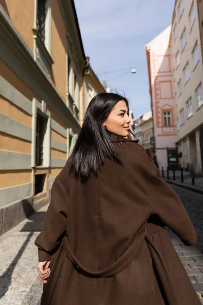 Vista lateral de mujer morena en abrigo caminando por la calle en Praga - foto de stock