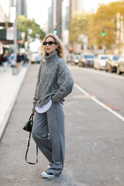 Full length of stylish woman in sunglasses and grey outfit holding handbag while posing on street of New York city — Stock Photo