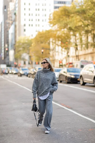 Comprimento total de mulher loira em óculos de sol e roupa na moda segurando bolsa enquanto caminhava na rua da cidade de Nova York — Fotografia de Stock