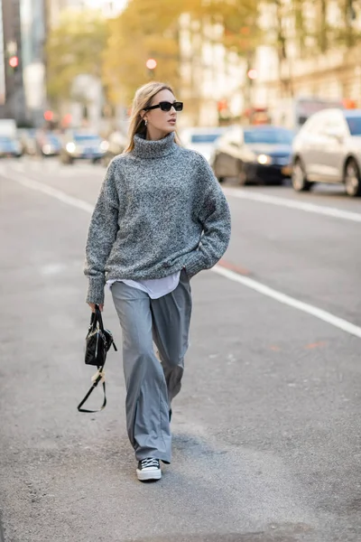 Piena lunghezza della donna bionda in occhiali da sole e vestito grigio che tiene la borsetta mentre cammina sulla strada di New York — Foto stock