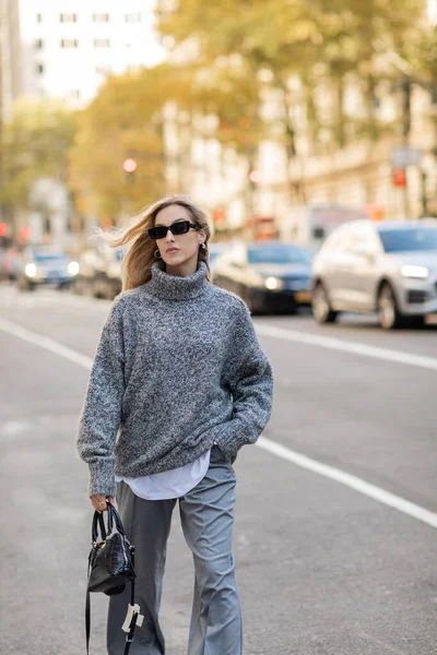 Blonde woman in sunglasses and grey outfit holding handbag while walking with hand in pocket on street of New York city — Stock Photo