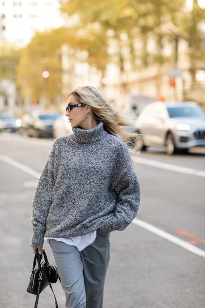 Jovem mulher em óculos de sol e roupa cinza segurando bolsa enquanto de pé com a mão no bolso na rua da cidade de Nova York — Fotografia de Stock