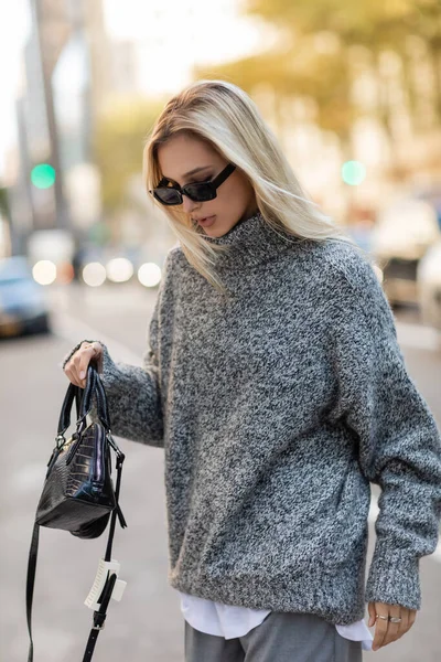 Young woman in sunglasses and grey sweater holding handbag while walking on street of New York city — Stock Photo