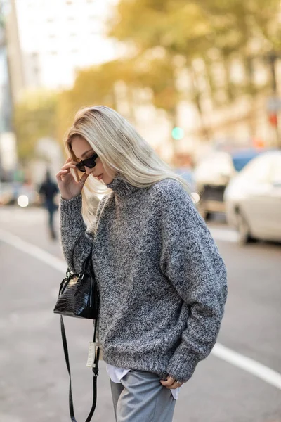 Mulher loira em camisola cinza segurando bolsa e ajustando óculos de sol na moda na rua da cidade de Nova York — Fotografia de Stock