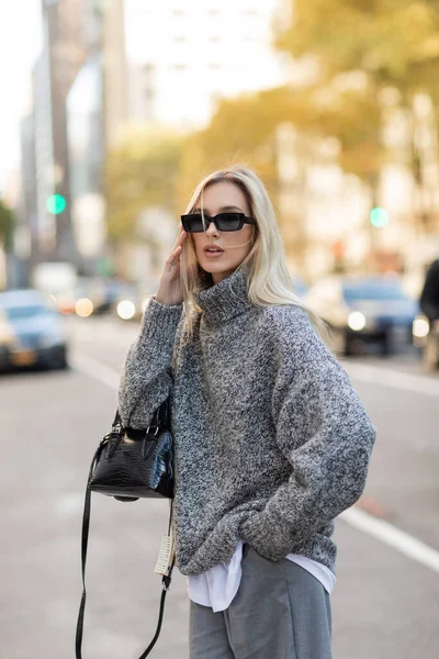 Blonde woman in grey sweater holding handbag and standing with hand in pocket on street of New York city — Stock Photo