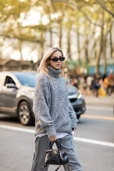 Mujer de moda en suéter gris y gafas de sol sosteniendo el bolso mientras camina por la calle de la ciudad de Nueva York - foto de stock