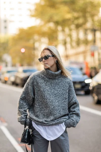 Стильная женщина в солнечных очках и сером костюме, держащая черную сумочку и стоя с руками в карманах на улице Нью-Йорка — стоковое фото