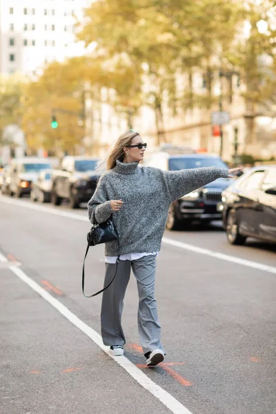 Piena lunghezza della donna alla moda in occhiali da sole e vestito grigio in possesso di borsetta e prendere un taxi sulla strada di New York — Foto stock