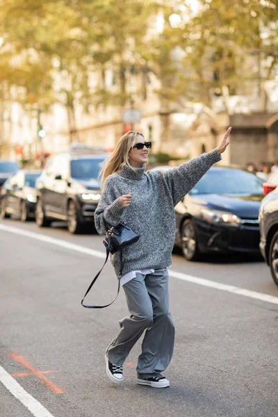 Comprimento total de mulher alegre em óculos de sol e roupa cinza segurando bolsa e pegar táxi na rua de Nova York — Fotografia de Stock