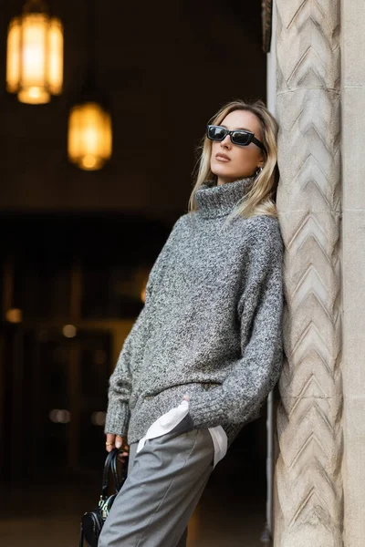 Mulher loira em óculos de sol e roupa cinza segurando bolsa enquanto está de pé com a mão no bolso perto do edifício em Nova York — Fotografia de Stock