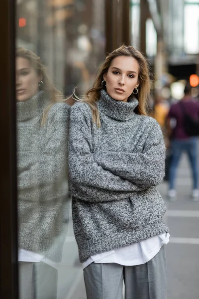Comprimento total da jovem mulher em roupas de inverno em pé com braços cruzados perto de vitrine em Nova York — Fotografia de Stock