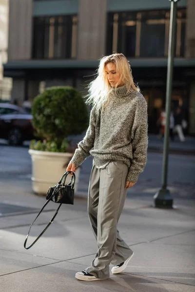 Full length of stylish young woman with blonde hair holding handbag while walking in New York city in winter — Stock Photo