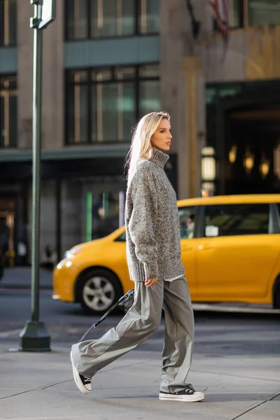 Longitud completa de mujer joven con estilo con el pelo rubio caminando cerca de taxi amarillo en Nueva York - foto de stock