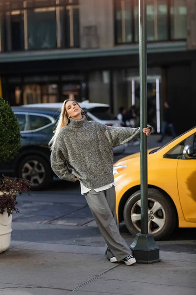 Longitud completa de la mujer joven con estilo en traje de invierno de moda posando cerca de poste de la calle y taxi amarillo en Nueva York - foto de stock