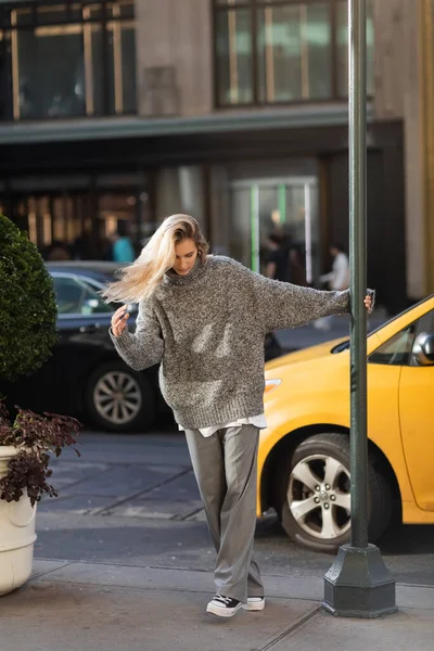Longitud completa de la mujer joven en traje de invierno elegante posando cerca de poste de la calle y taxi amarillo en Nueva York - foto de stock