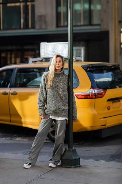 Longitud completa de la joven en traje de invierno gris posando cerca de poste de la calle y taxi amarillo en Nueva York - foto de stock