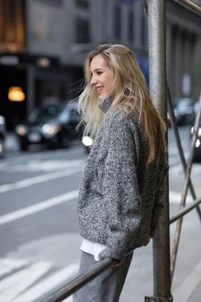 Mujer alegre en suéter de punto gris sonriendo en la calle urbana de la ciudad de Nueva York - foto de stock